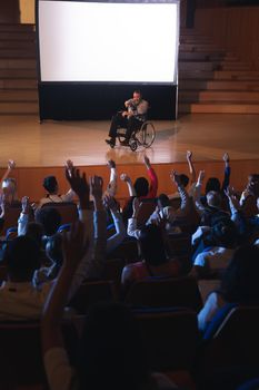 High view of Caucasian businessman sitting on a wheelchair and giving presentation to the audience while audience raising hand for asking question in auditorium