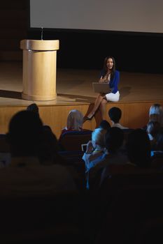 High view of beautiful businesswoman sitting at the side of the stage and having laptop in her hand in front of audience