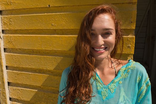 Portrait of gorgeous young Caucasian woman smiling and leaning standing against beach hut. She is looking at camera