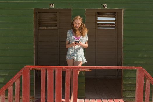 Front view of happy gorgeous young Caucasian woman using mobile phone at beach hut. She is smiling