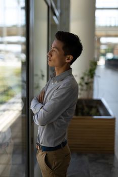 Side view of young Asian male executive standing with arms crossed in modern office. He is thinking