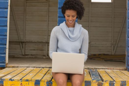 Front view of happy young African American woman using her laptop sitting on the beach hut