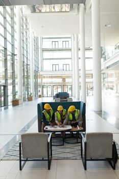  High angle view of young multi-ethnic business architect sitting and discussing over the blue print in lobby at office. They wear safety helmets