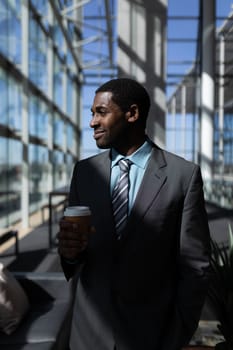 Front view of thoughtful young African-American businessman with coffee cup looking away in modern office. He is smiling