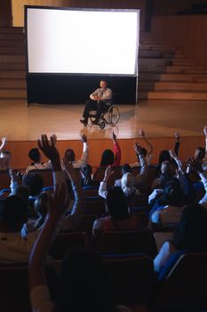 High view of Caucasian businessman sitting on a wheelchair and giving presentation to the audience while audience raising hand for asking question in auditorium