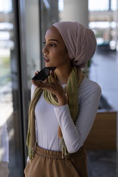 Front view of thoughtful beautiful youg mixed-race female executive talking on mobile phone standing in modern office