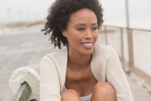 Front view of happy young African American woman sitting on bench at promenade. She is smiling