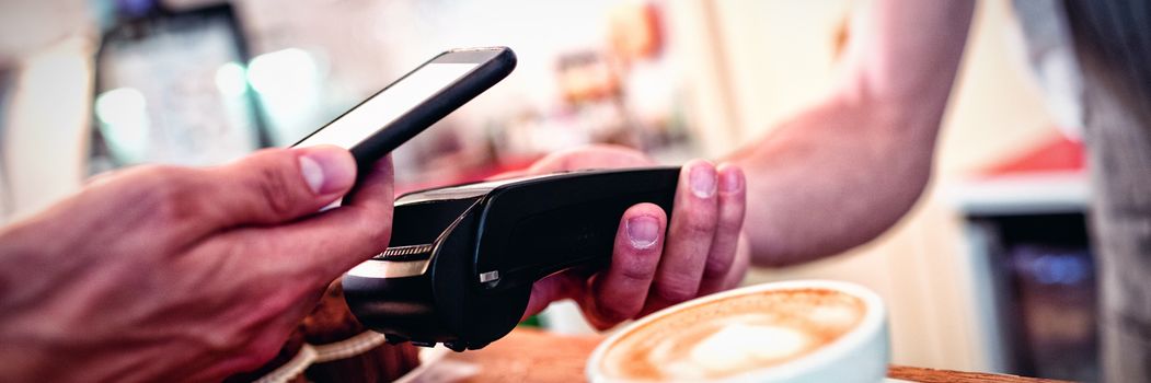 Close-up of customer with cellphone and male barista with card reader at counter in coffee shop