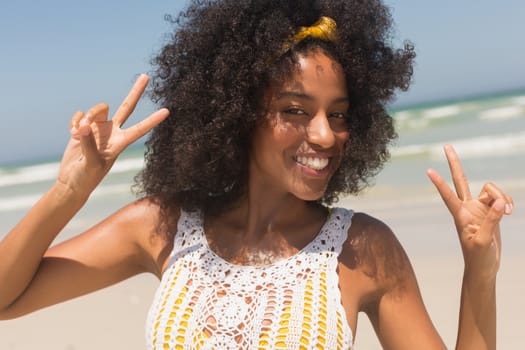 Front view of happy young African American woman showing victory sign standing on the beach