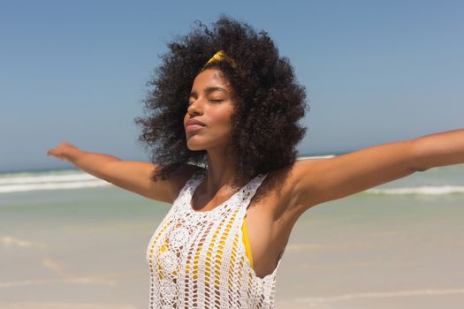 Side view of young African American woman with eyes closed and arms stretched out standing on beach in the sunshine. She is relaxed