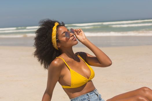 Side view of young African American woman in yellow bikini and sunglasses relaxing on beach in the sunshine. She is enjoying her holidays