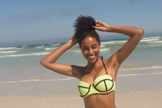 Portrait of young African American woman in green bikini and hands in hair looking at camera on the beach. She is smiling