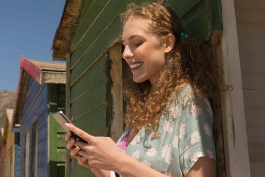 Side view of happy gorgeous young woman using mobile phone at beach hut. She is smiling