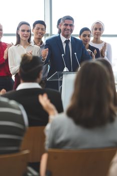 Front view of mixed race businessman standing on podium with diverse colleagues while he speaks at business seminar in office building