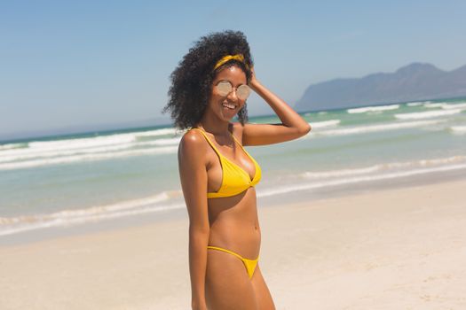 Portrait of happy young African American woman in yellow bikini and sunglasses standing on the beach. She is smiling and looking at camera