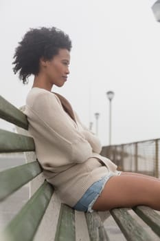 Side view of thoughtful young African American woman with arms crossed sitting on bench at promenade