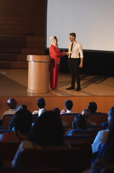 Front view of mixed race business colleague standing and discussing with each other in front of the audience in auditorium while shaking hand 