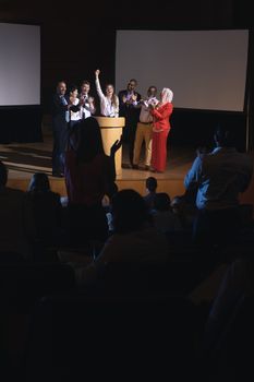 High view of happy Caucasian businesswoman standing at the stage of the auditorium with colleagues in front of audience