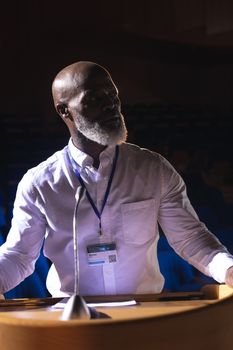 Side view of old African-American businessman looking and thinking while standing on stage in auditorium