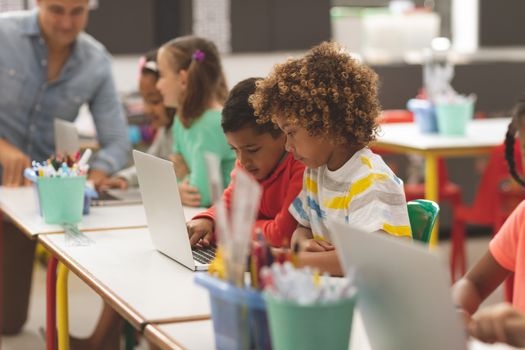 Side view of school kids using laptop in classroom while teacher teaching in background at school