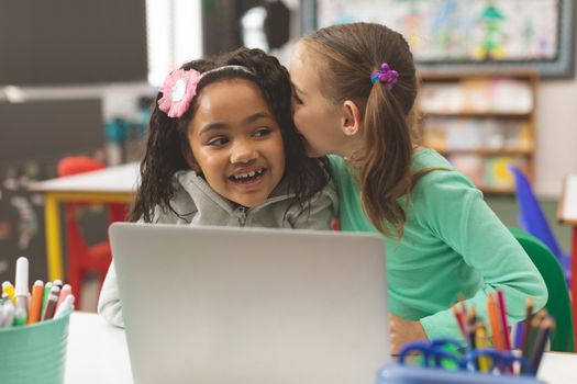 Front view of a Caucasian schoolgirl whispering a secret into friend ear while her friend laughing in classroom at school
