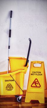 Yellow sign board with mop bucket on wooden floor against wall