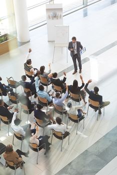 High angle view of group of diverse business people raising their hands to ask questions to handsome middle-aged Caucasian businessman at conference.