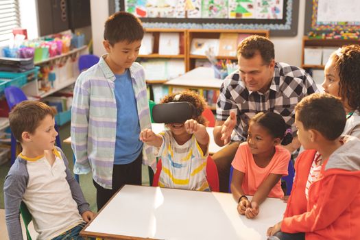 Front view of a schoolboy using virtual reality headset with his classmate and teacher at school