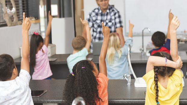 Rear view of school kids raising hand while teacher asking question in classroom at school