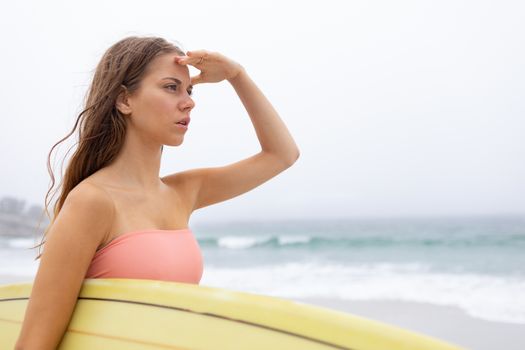 Rear view beautiful youngh Caucasian female surfer standing with her surfboard on the beach. She is looking away