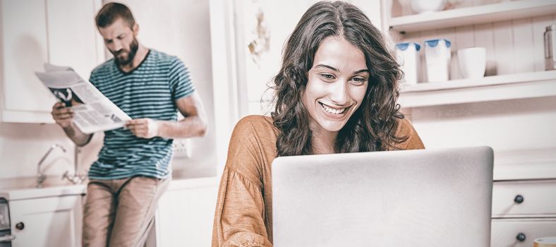 Woman using laptop while man reading newspaper in background at kitchen