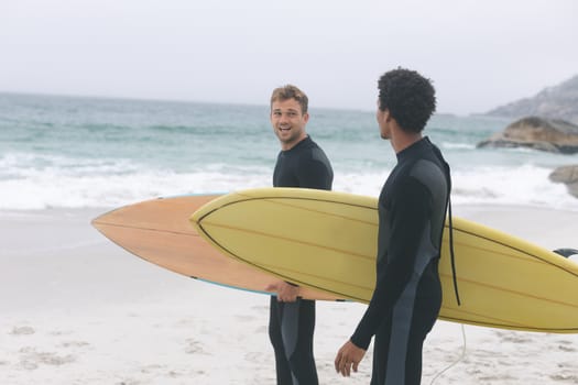 Front view of young multi-ethnic male friends interacting with each other on beach while holding surfboards. They are smiling