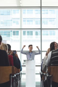 Front view of young Asian businessman cheering during seminar in front of business professionals sitting in office building