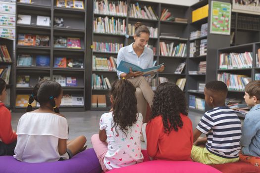 Front view of a teacher reading a story in a library to school kids sitting over big colored cushions in foreground