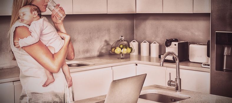 Young woman drinking water while carrying baby in kitchen at home