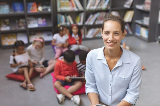 Overhead view of a happy teacher looking and smilling at the camera while school kids relaxing themselves over digital tablet on cushions in background