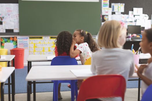 Rear view of schoolgirl whispering into her friend ear in classroom at school