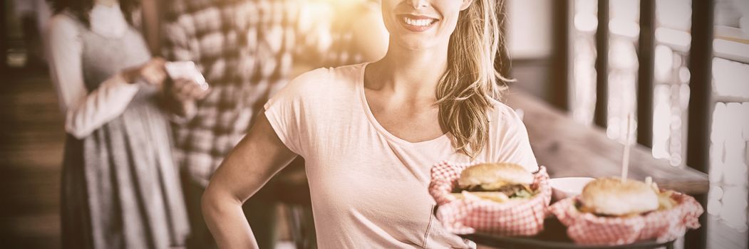 Portrait of smiling young waitress serving burger with customers in background at restaurant