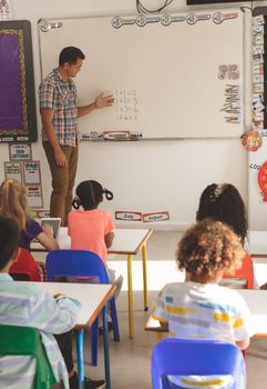 Side view of teacher teaching mathematics at his school kids sitting on their colored chairs in classroom at school