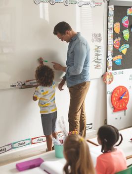 Rear view of a Caucasian teacher teatching something to a metis school boy on a whiteboard with school girls looking at him at the first row against drawing in background