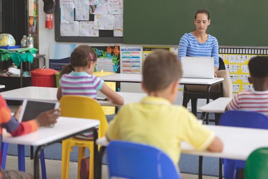 Front view of a teacher using laptop in classroom while school kids studying in classroom at school