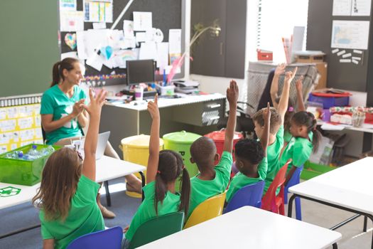 Front view of caucasian teacher discussing about green energy and recycle while multi ethnic students raising their hands at desk in classroom
