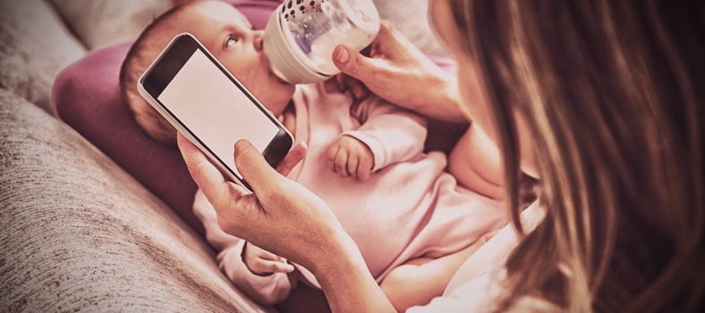 Mother using mobile phone while feeding her baby with milk bottle at home