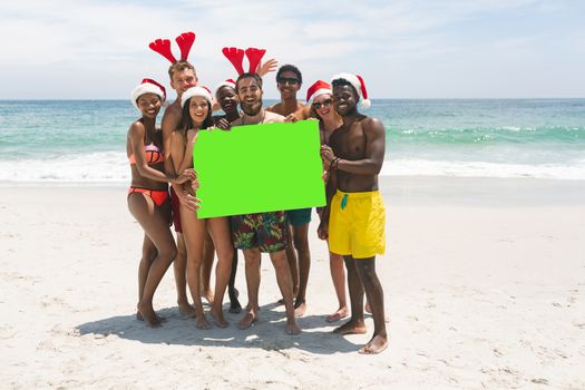 Front view of happy multi-ethnic group of friends with Christmas hat holding a empty green placard at beach on a sunny day. They are smiling and looking at camera
