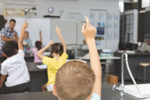 Rear view of school kids raising hand in classroom at school with teacher in background