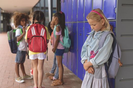 Side view of a sad schoolgirl standing in corridor while others school kids nteracting with each other at school
