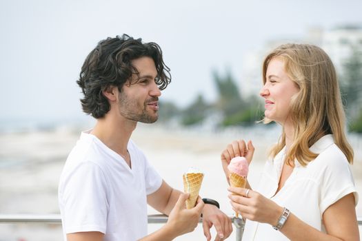 Side view of happy young Caucasian couple standing at promenade while having ice cream cone. They are smiling