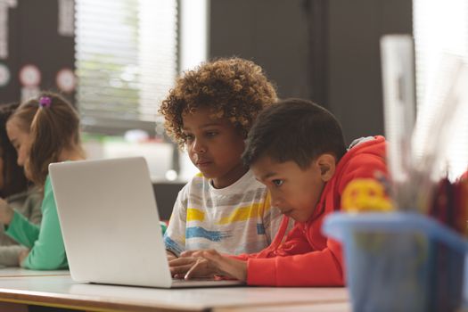 Front view of two school kids working on one laptop in classroom against school girl in background