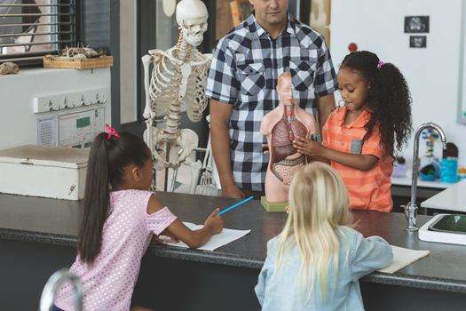 Front view of a teacher and a schoolgirl behind a desk showing to others school kids a dummy skeleton in classroom at school