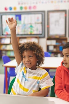 Front view of mixed-race schoolboy raising his hand in with his friend next to him in classroom at school